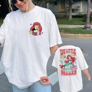 May include: White t-shirt featuring a colorful graphic of Ariel from The Little Mermaid. The front has a small image of Ariel with a starfish. The back of the shirt has a larger design with Ariel, the text "THE LITTLE MERMAID", and other sea creatures.