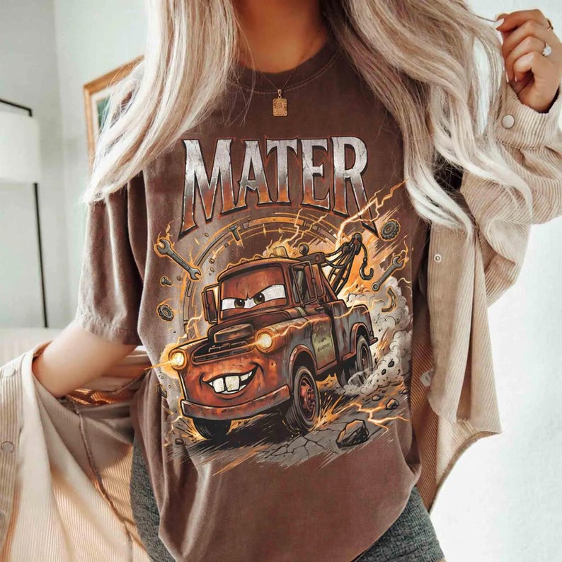 Może przedstawiać: Brązowa koszulka z napisem "MATER" metaliczną czcionką nad kresk&oacute;wkowym wizerunkiem Złomka z filmu Auta. Grafika zawiera błyskawice, narzędzia i wz&oacute;r prędkościomierza.