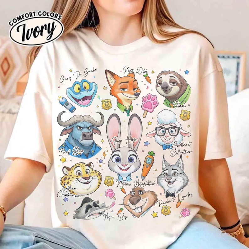 K&ouml;nnte beinhalten: Elfenbeinfarbenes T-Shirt mit Zeichentrickfiguren aus einem Film. Das Design umfasst eine Frosch, einen Fuchs, ein Faultier, ein Kaninchen, ein Schaf, einen Geparden und andere Charaktere. Das Shirt tr&auml;gt die Aufschrift "COMFORT COLORS" und "Ivory".
