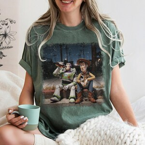 Puede incluir: Camiseta verde con un gr&aacute;fico de Buzz Lightyear y Woody de Toy Story. La imagen muestra a los personajes sentados en una acera por la noche. Buzz est&aacute; bebiendo de una botella. La camiseta tiene un aspecto vintage.