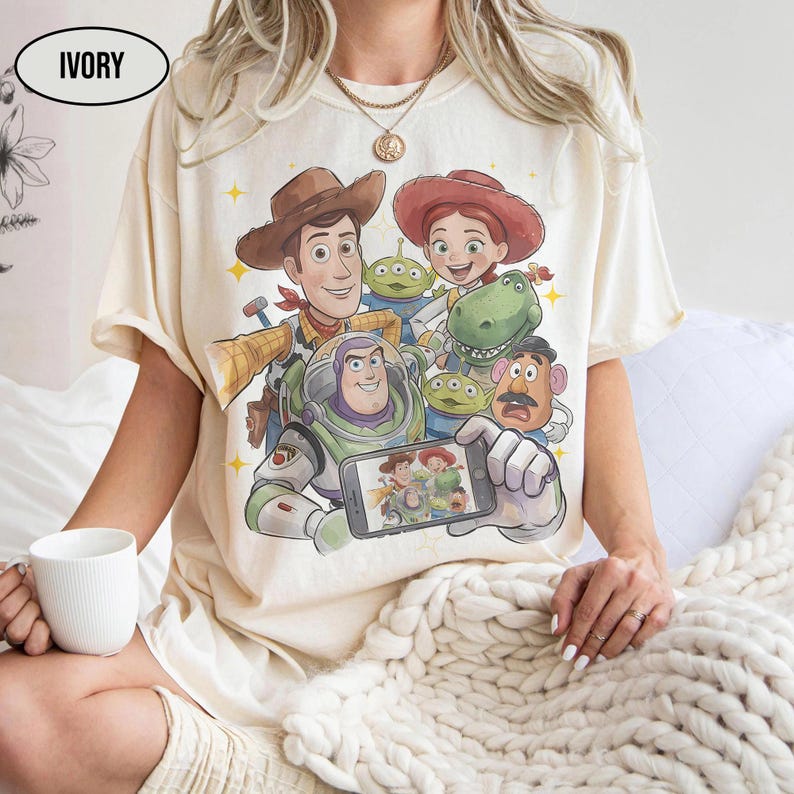 May include: Ivory-colored t-shirt featuring a colorful graphic of characters from a popular animated movie. The design includes Woody, Buzz Lightyear, Jessie, and other characters. The shirt has short sleeves and a crew neck.