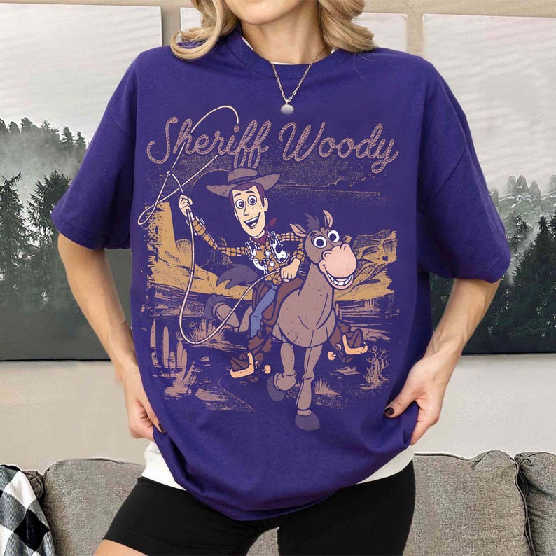 May include: A purple t-shirt featuring a graphic of Sheriff Woody from Toy Story riding a horse. The text "Sheriff Woody" is arched above the image. The design is in shades of brown, tan, and white.