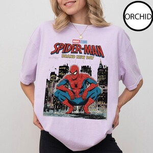 May include: A light purple t-shirt featuring a Marvel Studios Spider-Man graphic. The graphic shows Spider-Man crouching in front of a cityscape, with the words "SPIDER-MAN BRAND NEW DAY" in red and blue.