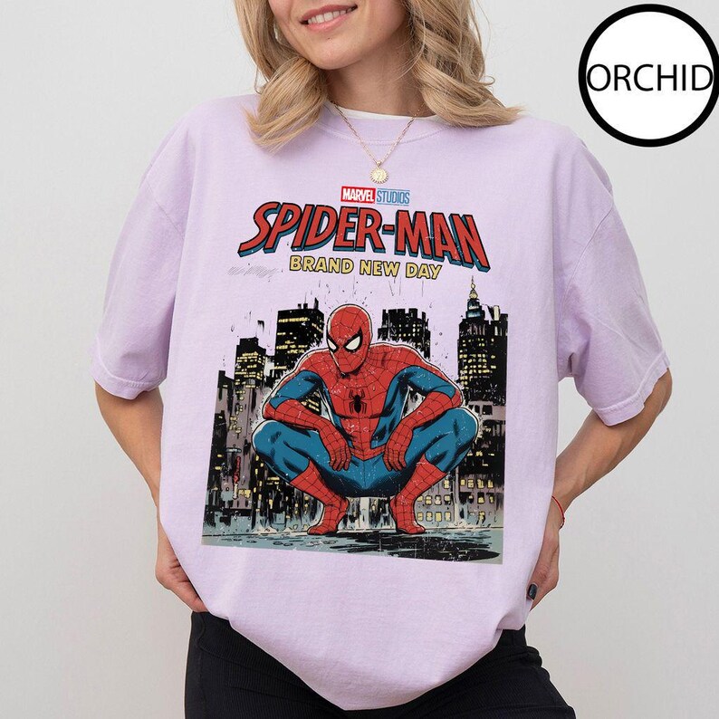 May include: A light purple t-shirt featuring a Marvel Studios Spider-Man graphic. The graphic shows Spider-Man crouching in front of a cityscape, with the words "SPIDER-MAN BRAND NEW DAY" in red and blue.