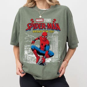 Pode incluir: Camiseta verde s&aacute;lvia com um gr&aacute;fico do Homem-Aranha da Marvel Studios. O design inclui o logotipo do Homem-Aranha, as palavras "Spider-Man Brand New Day" e uma ilustra&ccedil;&atilde;o do Homem-Aranha em uma pose din&acirc;mica contra um cen&aacute;rio urbano.