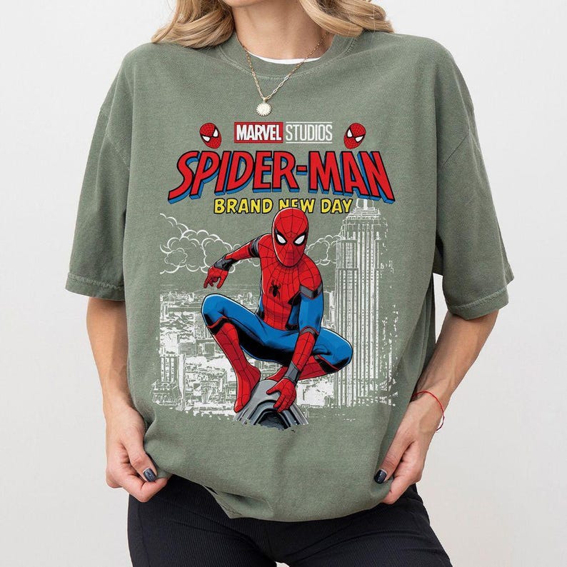 Pode incluir: Camiseta verde s&aacute;lvia com um gr&aacute;fico do Homem-Aranha da Marvel Studios. O design inclui o logotipo do Homem-Aranha, as palavras "Spider-Man Brand New Day" e uma ilustra&ccedil;&atilde;o do Homem-Aranha em uma pose din&acirc;mica contra um cen&aacute;rio urbano.