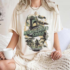 K&ouml;nnte beinhalten: Cremefarbenes T-Shirt mit einer Grafik aus der Star Wars-Serie "The Mandalorian and Grogu". Das Design zeigt den Helm des Mandalorianers, Grogu und den Satz "THIS IS THE WAY".
