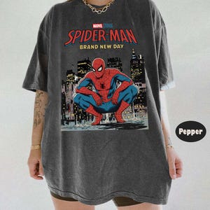 May include: Dark gray t-shirt featuring a graphic of Spider-Man in a crouching pose against a city skyline. The shirt has the text "MARVEL STUDIOS SPIDER-MAN BRAND NEW DAY" in red and blue.