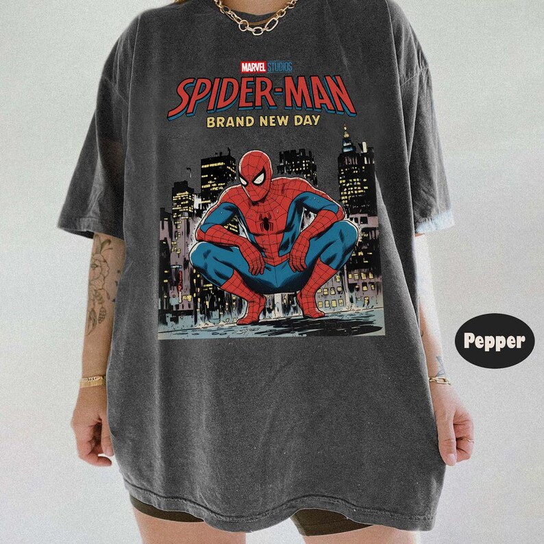 May include: Dark gray t-shirt featuring a graphic of Spider-Man in a crouching pose against a city skyline. The shirt has the text "MARVEL STUDIOS SPIDER-MAN BRAND NEW DAY" in red and blue.