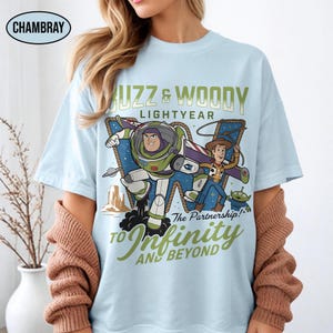 以下が含まれることがあります： トイ・ストーリーのバズ・ライトイヤーとウッディが描かれた水色のTシャツ。「Buzz & Woody Lightyear」と「To Infinity and Beyond!」の文字が緑と金でデザインされています。カジュアルウェアです。