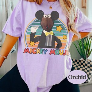May include: A light purple t-shirt featuring a cartoon Mickey Mouse character in a tuxedo, set against a striped background with stars. The text "Mickey Mouse" is displayed in colorful letters. The word "Orchid" is printed on the bottom right.