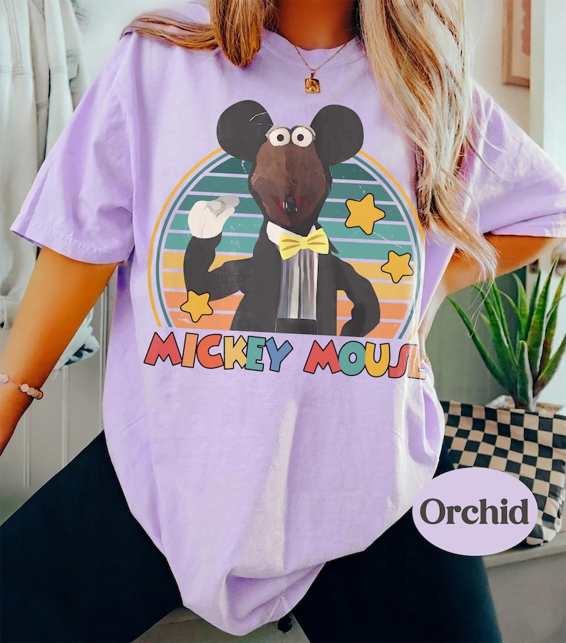 May include: A light purple t-shirt featuring a cartoon Mickey Mouse character in a tuxedo, set against a striped background with stars. The text "Mickey Mouse" is displayed in colorful letters. The word "Orchid" is printed on the bottom right.