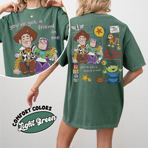 Op de afbeelding: Lichtgroen T-shirt met een cartoon illustratie van Woody en Buzz Lightyear en de tekst "you've got a friend in me." De achterkant van het shirt heeft extra Toy Story graphics, waaronder Slinky Dog en het Pizza Planet logo.
