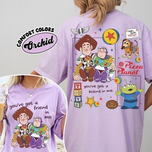 Op de afbeelding: Een lichtpaars Comfort Colors T-shirt met stripfiguren Woody en Buzz Lightyear. Het shirt heeft de tekst "you've got a friend in me" en "Pizza Planet" erop gedrukt. Het shirt heeft ook het woord "Orchid" erop gedrukt.