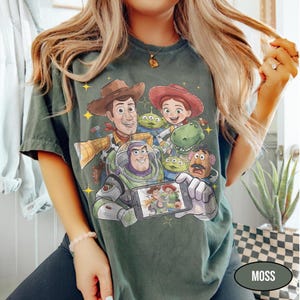May include: Moss green t-shirt featuring a colorful graphic of Toy Story characters, including Woody, Buzz Lightyear, and Jessie. The design includes the characters posing for a selfie. The shirt is a relaxed fit.