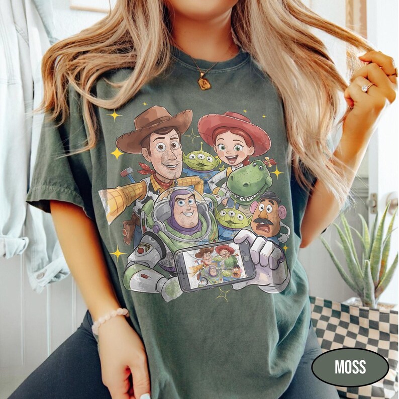 May include: Moss green t-shirt featuring a colorful graphic of Toy Story characters, including Woody, Buzz Lightyear, and Jessie. The design includes the characters posing for a selfie. The shirt is a relaxed fit.