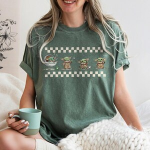 May include: Green t-shirt featuring a graphic of Grogu in various poses, set against a white and black checkered pattern. The design includes Grogu in a pod, with a frog, and holding a cup. The shirt has rolled-up sleeves.