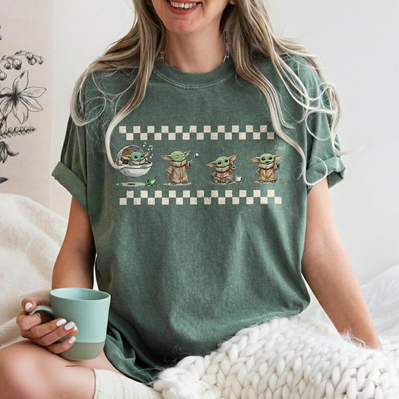 May include: Green t-shirt featuring a graphic of Grogu in various poses, set against a white and black checkered pattern. The design includes Grogu in a pod, with a frog, and holding a cup. The shirt has rolled-up sleeves.
