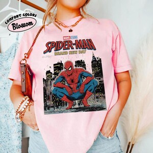 May include: A light pink t-shirt featuring a Marvel Studios Spider-Man graphic. The design includes Spider-Man in a crouching pose with a cityscape background and the words "Spider-Man Brand New Day". The shirt also has the words "Comfort Colors Blossom".