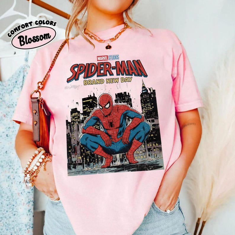 May include: A light pink t-shirt featuring a Marvel Studios Spider-Man graphic. The design includes Spider-Man in a crouching pose with a cityscape background and the words "Spider-Man Brand New Day". The shirt also has the words "Comfort Colors Blossom".