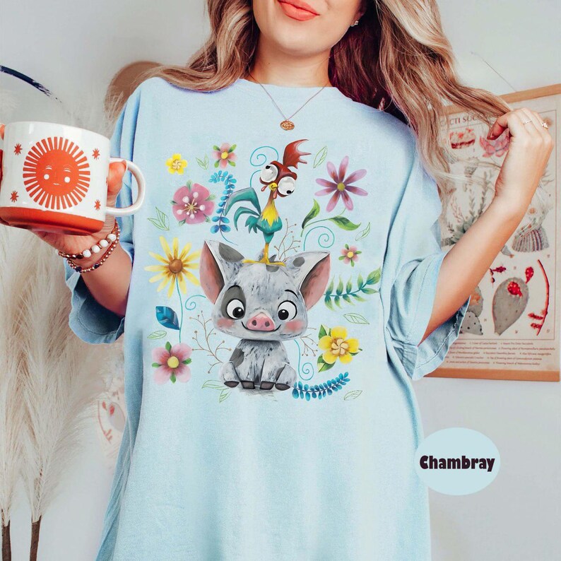 May include: Pale blue t-shirt with a cartoon illustration of a pig, rooster, and flowers. The shirt has a loose fit and is made of a soft fabric. The word "Chambray" is printed on the bottom right.