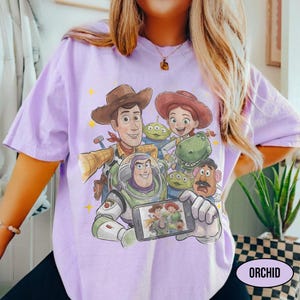 May include: A light purple t-shirt featuring a graphic of characters from the Toy Story movie. The design includes Woody, Buzz Lightyear, Jessie, and other characters. The word "ORCHID" is printed on the bottom right.