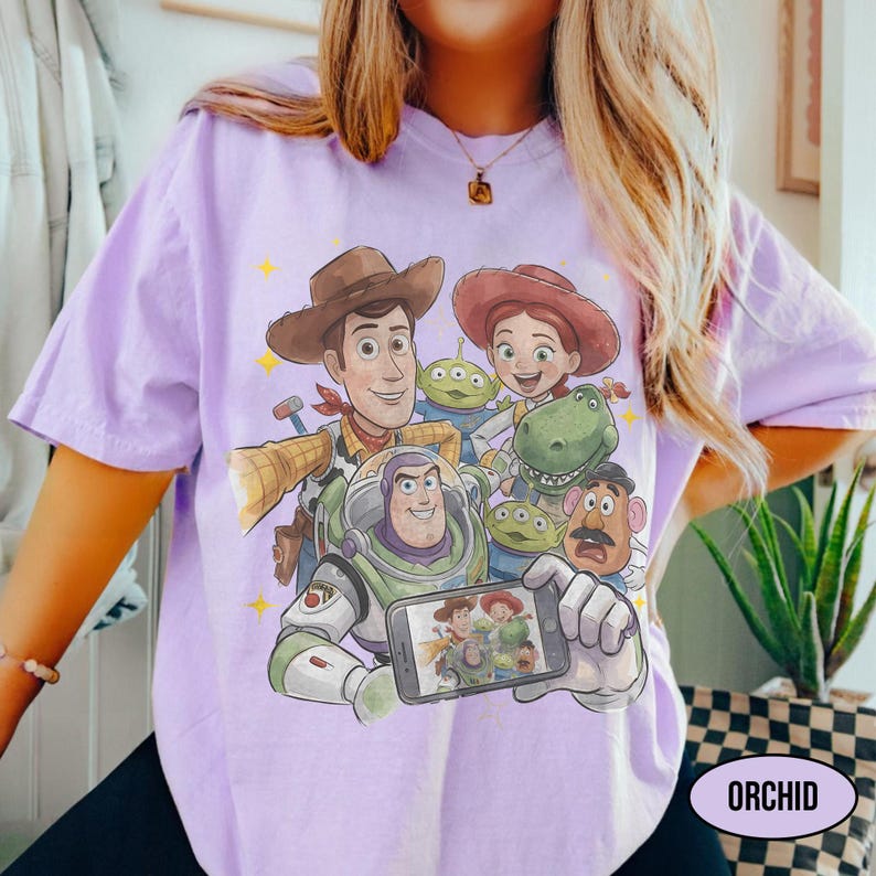 May include: A light purple t-shirt featuring a graphic of characters from the Toy Story movie. The design includes Woody, Buzz Lightyear, Jessie, and other characters. The word "ORCHID" is printed on the bottom right.