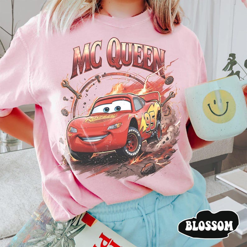 May include: A light pink t-shirt featuring a graphic of Lightning McQueen from the movie Cars. The design includes the text "MC QUEEN" above the car and the number "95" on the side. The word "BLOSSOM" is printed at the bottom.
