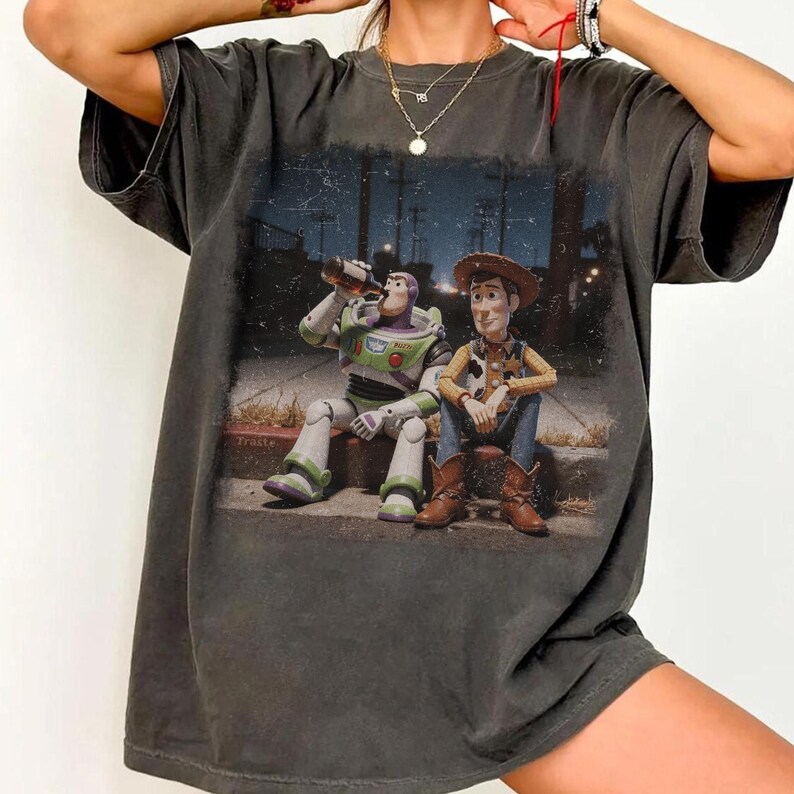 Puede incluir: Camiseta gris oscuro con un gr&aacute;fico de Buzz Lightyear bebiendo de una botella junto a Woody. La imagen tiene un aspecto vintage y desgastado. Los personajes est&aacute;n sentados en una acera con un fondo nocturno.