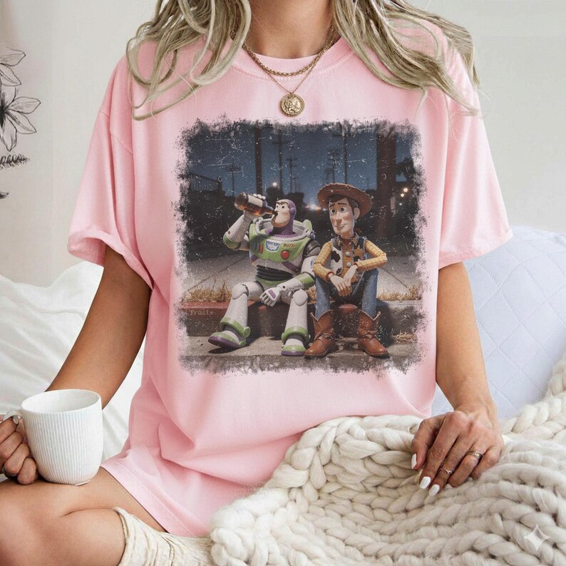 Puede incluir: Camiseta rosa claro con un estampado gr&aacute;fico de Buzz Lightyear y Woody de Toy Story. La imagen muestra a los personajes sentados en una acera, con Buzz bebiendo de una botella. La camiseta tiene mangas cortas y cuello redondo.