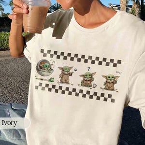 May include: Ivory-colored t-shirt featuring a graphic of a character from a popular science fiction series. The design includes the character in various poses, with a frog, musical notes, and the word "Cool!" printed on the shirt.