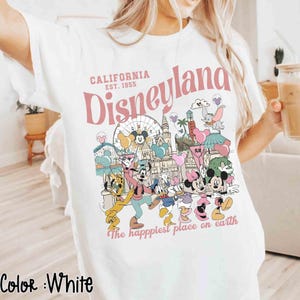 May include: White t-shirt with a Disneyland graphic in pink and other colors. The design includes the words "California Disneyland Est. 1955" and various Disney characters. The phrase "The happiest place on earth" is also printed on the shirt.
