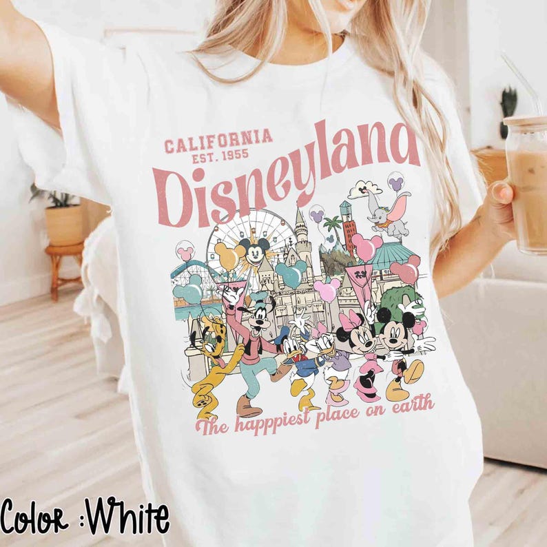 May include: White t-shirt with a Disneyland graphic in pink and other colors. The design includes the words "California Disneyland Est. 1955" and various Disney characters. The phrase "The happiest place on earth" is also printed on the shirt.