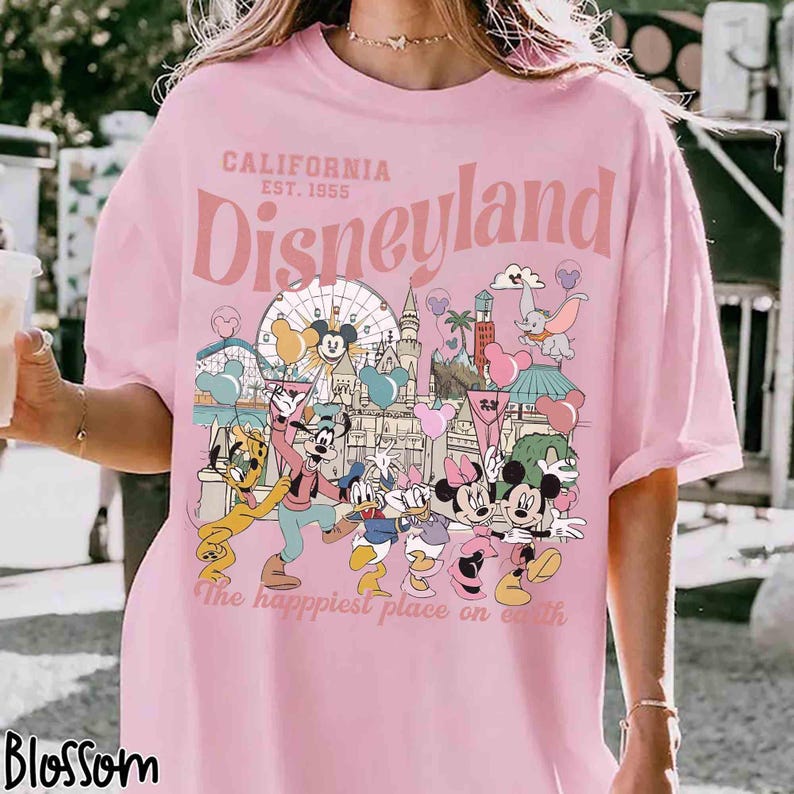 May include: A pink t-shirt featuring a Disneyland graphic. The design includes the text "California Est. 1955 Disneyland" and cartoon characters such as Mickey and Minnie Mouse. The phrase "The happiest place on earth" is also included in the design.