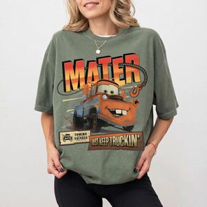 Puede incluir: Camiseta verde de manga corta con un dise&ntilde;o de Mate de la pel&iacute;cula Cars. El dise&ntilde;o incluye la palabra "MATER" en naranja y amarillo, y la frase "JUST KEEP TRUCKIN'". La camiseta tambi&eacute;n tiene un peque&ntilde;o logo de "Towing & Salvage".