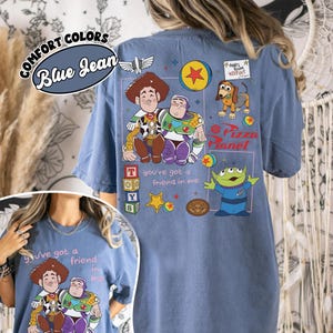 Op de afbeelding: Blauw spijkerbroekkleurig T-shirt met de tekenfilmfiguren Woody en Buzz Lightyear. Het shirt heeft de tekst "You've got a friend in me" en "Pizza Planet". Het shirt heeft ook de tekst "Andy's Room Keep Out".