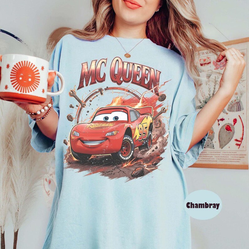 May include: Light blue t-shirt featuring a graphic of Lightning McQueen from the movie Cars. The design includes the text "MC QUEEN" and the number "95" on the car. A red and white mug with a sun design is held in the image.