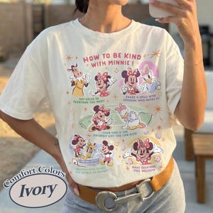 Peut inclure: T-shirt ivoire avec un motif de Minnie Mouse et ses amis. Le texte "HOW TO BE KIND WITH MINNIE!" est pr&eacute;sent, ainsi que des phrases comme "Dites bonjour &agrave; tout le monde !" et "Faites en sorte que tout le monde se sente bienvenu."