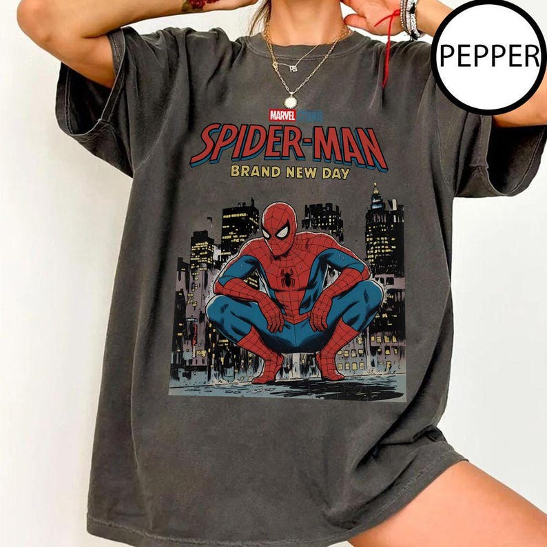 May include: Dark gray t-shirt featuring a graphic of Spider-Man in a crouched pose against a cityscape backdrop. The shirt displays the text "SPIDER-MAN BRAND NEW DAY" in red and blue, with the Marvel logo above.