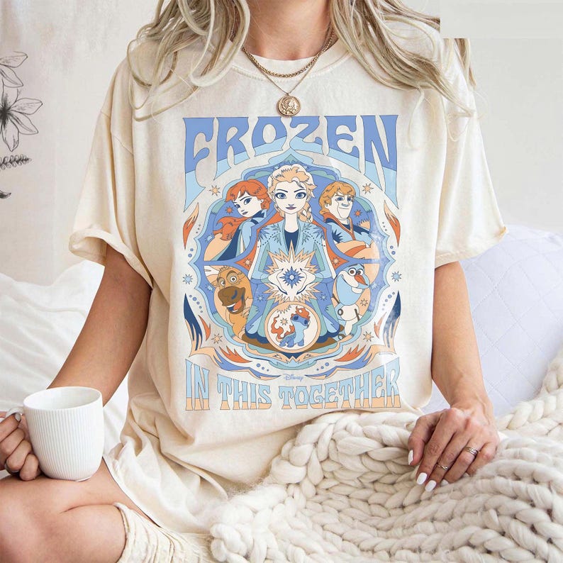 May include: Cream-colored t-shirt featuring a graphic from the Disney movie Frozen. The design includes the word "FROZEN" in blue, and the phrase "IN THIS TOGETHER". The graphic showcases characters Anna, Elsa, Kristoff, and Olaf.