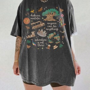 Pode incluir: T-shirt cinzenta escura com um gr&aacute;fico colorido. O gr&aacute;fico inclui as palavras "hakuna matata", "anyone can be anything" e "adventure is out there", juntamente com ilustra&ccedil;&otilde;es de uma &aacute;rvore, animais e um autocarro.