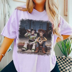 Puede incluir: Camiseta de color lila claro con un gr&aacute;fico desgastado de Buzz Lightyear y Woody de Toy Story. Buzz est&aacute; bebiendo de una botella, mientras que Woody est&aacute; sentado a su lado. El gr&aacute;fico est&aacute; ambientado en una escena callejera nocturna.