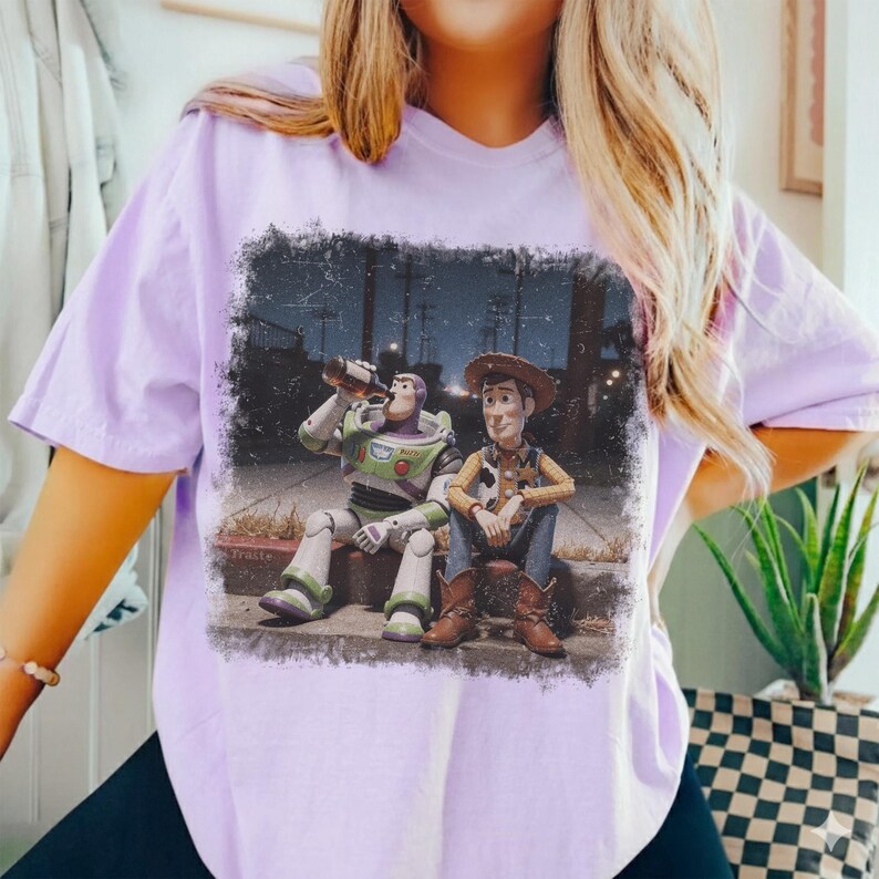 Puede incluir: Camiseta de color lila claro con un gr&aacute;fico desgastado de Buzz Lightyear y Woody de Toy Story. Buzz est&aacute; bebiendo de una botella, mientras que Woody est&aacute; sentado a su lado. El gr&aacute;fico est&aacute; ambientado en una escena callejera nocturna.