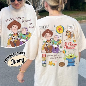 Op de afbeelding: Ivoor T-shirt met een afbeelding van Woody en Buzz Lightyear en de tekst "you've got a friend in me". De achterkant van het shirt toont andere Toy Story-personages en het Pizza Planet-logo.