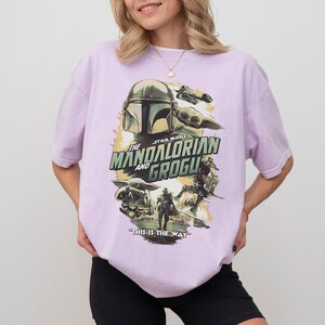 K&ouml;nnte beinhalten: Ein helllila T-Shirt mit einer Grafik aus der Star Wars-Serie "The Mandalorian and Grogu". Das Design zeigt Bilder von Mandalorian, Grogu und anderen Charakteren mit dem Satz "This is the Way."