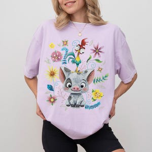 May include: A lilac t-shirt with a vibrant graphic of a pig, rooster, and floral elements. The pig is grey and white, the rooster is yellow and red, and the flowers are various colours. The shirt has a relaxed fit.