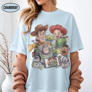 May include: Light blue t-shirt featuring a group of cartoon characters, including a cowboy, a space ranger, and a cowgirl, taking a selfie. The word "CHAMBRAY" is printed on the top left of the shirt. The characters are surrounded by yellow stars.