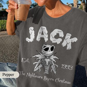 Puede incluir: Una camiseta gris carb&oacute;n con el texto blanco JACK en una fuente que gotea sobre un gr&aacute;fico de un personaje esqueleto envuelto en vendas. Debajo del personaje, el texto dice Est. 1993 y The Nightmare Before Christmas en letras blancas.