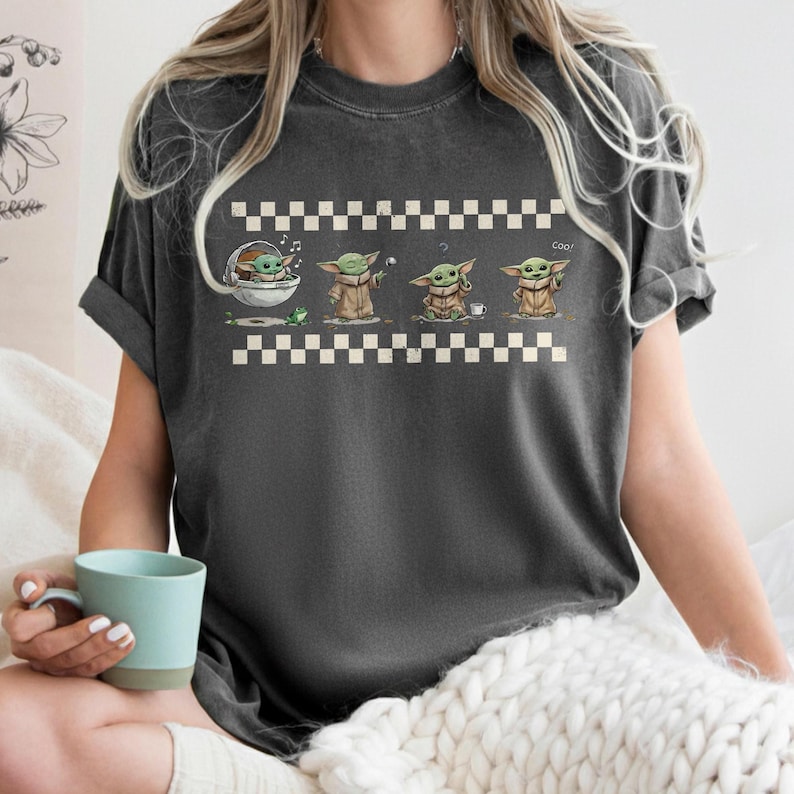May include: Dark gray t-shirt featuring a cartoon graphic of a character in various poses. The graphic includes a frog, a cup, and the word "Cool". The design is set against a checkered pattern.