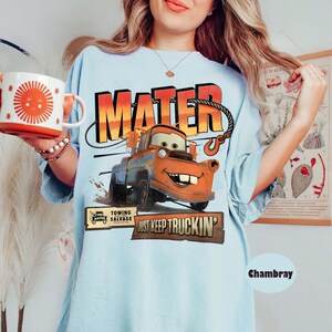 Puede incluir: Camiseta azul claro con un dise&ntilde;o de Mate de la pel&iacute;cula Cars, con la palabra "MATER" en naranja y amarillo. La camiseta tambi&eacute;n tiene el texto "JUST KEEP TRUCKIN'" y una taza con un sol sonriente.